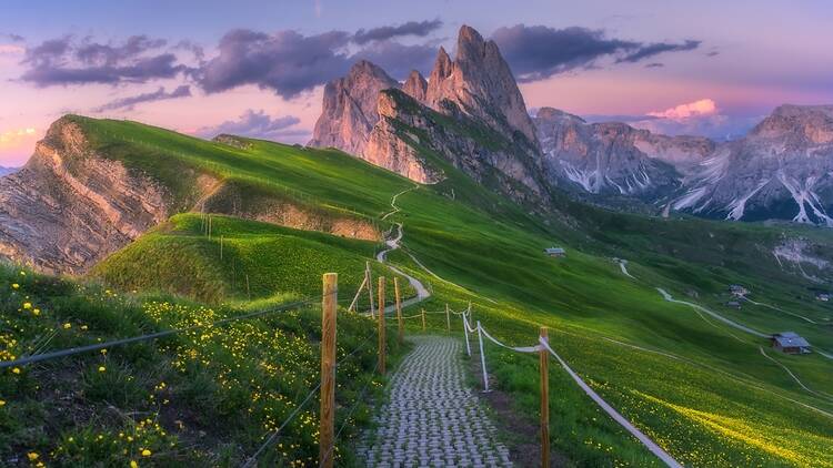Seceda ridgeline and Odle group at sunrise: stone path, green slopes, and pastel clouds
